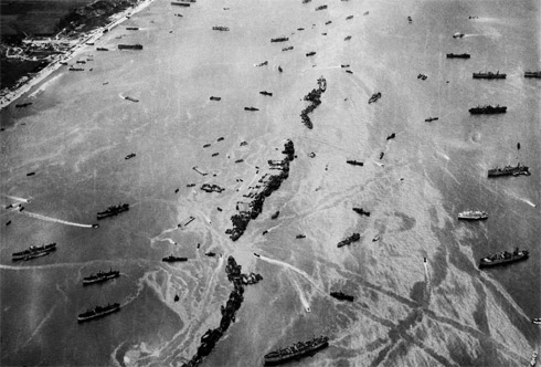 Thirteen liberty ships, deliberately scuttled to form a breakwater for invasion vessels landing on the Normandy beachhead lie in line off the beach, shielding the ships in shore. The artificial harbor engineering installation which was prefabricated and towed across the Channel. 1944 photo.